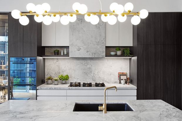 Contemporary kitchen with a large stone island and integrated sink, dark full-height cabinetry, white upper cabinets, and a matching stone splashback and rangehood surround. A sculptural brass-and-globe pendant light spans the room, with a glass-front beverage or display cabinet at left.