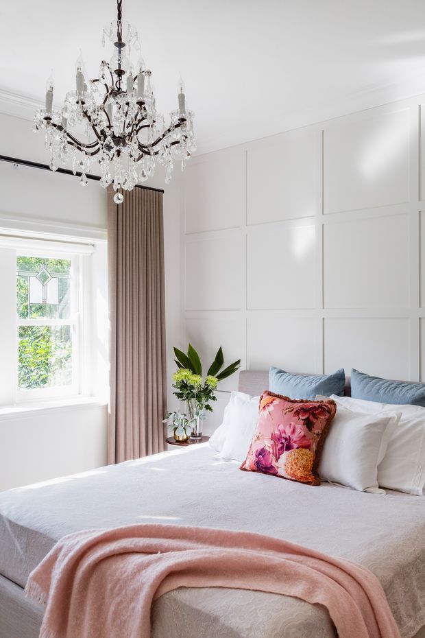 A bright, elegant bedroom with a large upholstered bed dressed in white bedding, layered blue and white pillows, and a floral pink accent cushion. A pale pink throw drapes across the bed. The room features white decorative wall panel moulding, a tall window with soft taupe curtains, and an ornate crystal chandelier. A small bedside table holds a leafy floral arrangement.