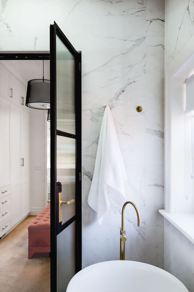 A refined bathroom with full-height white marble-look wall cladding featuring grey veining, a freestanding white bathtub, and a brushed gold floor-mounted tap. A black-framed ribbed-glass door opens directly onto a bedroom or dressing area with white built-in cabinetry and a blush upholstered bench. The space is bright and luxurious with strong black, white, marble, and gold detailing.