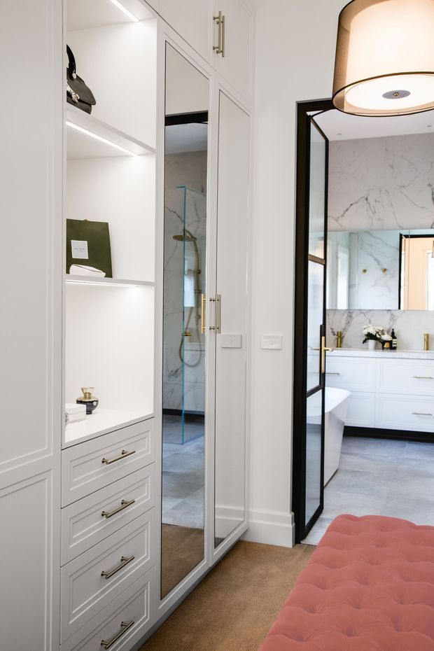 A refined dressing area opening directly into a luxurious marble bathroom. The foreground features built-in white painted cabinetry with mirrored wardrobe doors, brass hardware, illuminated open shelving, and drawers. A black-framed glass bathroom door reveals a walk-in shower and a white marble vanity with integrated cabinetry and brass fittings. A blush pink upholstered bench and oversized pendant light add colour and softness.