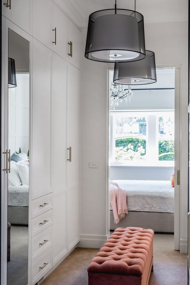 A refined bedroom dressing area or wardrobe passage with full-height white paneled built-in cabinetry, brass-toned hardware, mirrored wardrobe doors, and two large grey drum pendant lights. A blush pink tufted bench sits on a neutral carpet, while a bright bedroom with a bay window, bed, chandelier, and soft furnishings is visible beyond the opening.