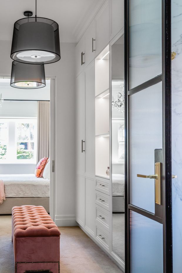 A refined bedroom entry and wardrobe area with a paneled white built-in storage wall, mirrored doors, drawers, and integrated lighting. A black-framed fluted-glass door with brass hardware occupies the foreground, while the adjoining bedroom contains a bed, upholstered blush-pink bench, and large pendant lights. The space is bright and elegant with detailed wall mouldings and soft neutral finishes.