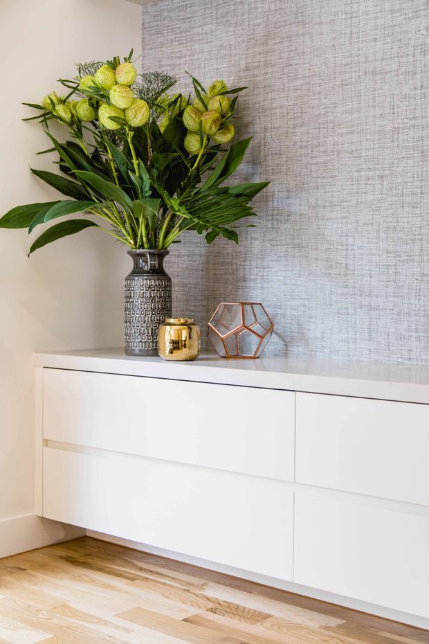 A bright interior vignette featuring a sleek white wall-mounted sideboard with handleless drawer fronts, positioned against a textured light grey wallpapered wall. A tall patterned metallic vase filled with lush green foliage and pale green flowers sits on the cabinet alongside a small gold vessel and geometric copper-toned ornament. Light timber flooring is visible below.