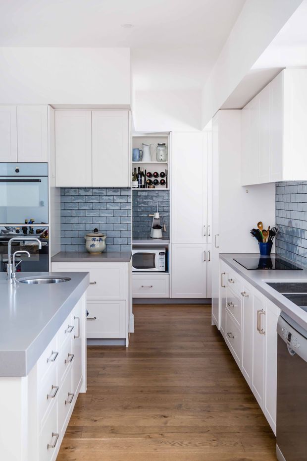 Bright galley-style kitchen with white shaker cabinetry, a central island with integrated sink, stainless-steel appliances, grey-blue subway tile splashbacks, open shelving, and warm timber flooring. The space has a clean, traditional-inspired fit-out with abundant built-in storage.