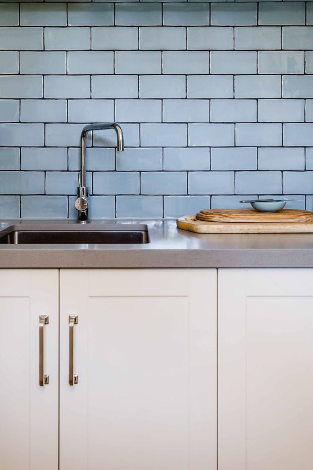 Close-up view of a compact kitchen sink area featuring pale blue rectangular subway tile backsplash with dark grout, a stainless-steel undermount sink and gooseneck faucet, a grey countertop, and white shaker-style cabinetry with slim metal pulls. A wooden cutting board and small bowl sit on the benchtop.