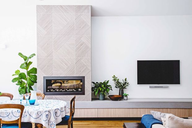 Bright contemporary living room with a wall-mounted television, long built-in timber media console, patterned feature wall surrounding a fireplace, indoor plants, timber flooring, and a dining table with a patterned tablecloth in the foreground.