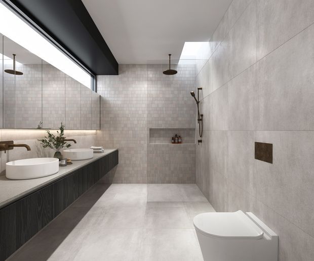Contemporary minimalist bathroom with a walk-in shower, wall-hung toilet, double vessel basins, and dark timber-look vanity cabinetry. Large-format pale grey wall and floor tiles are paired with a small square tiled feature wall, a recessed shower niche, black-framed glazing, and a skylight. The space has a clean, refined, softly illuminated material palette.