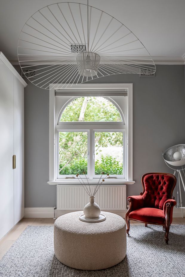 A refined sitting room with grey walls, white classical trim, a large arched-top sash window, and a striking oversized wireframe ceiling pendant. The room features a deep red tufted vintage armchair, a rounded textured cream ottoman, a pale rug, and a small sculptural side table with a floor lamp. Natural greenery is visible outside the window.