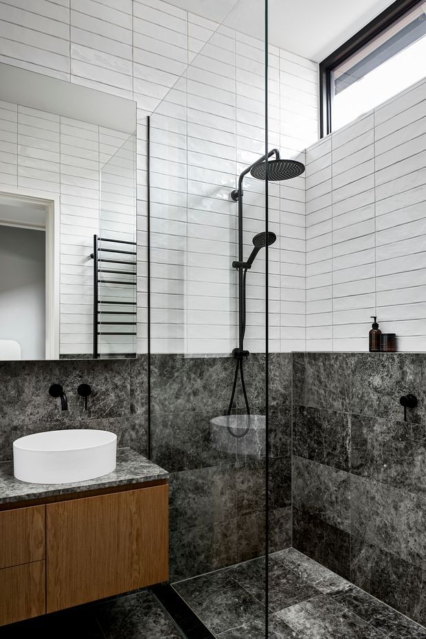 A contemporary bathroom featuring a frameless glass walk-in shower with matte black rain and hand shower fixtures, white rectangular wall tiles, dark veined stone wall and floor tiles, and a floating timber vanity with a white round vessel basin. A black heated towel rail and high window complete the bright, refined space.