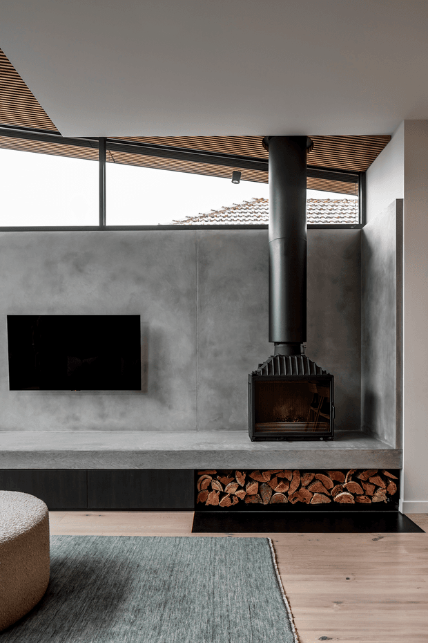A contemporary living room centred on a black freestanding wood-burning fireplace with a large cylindrical flue. The fireplace is integrated into a broad grey concrete-look feature wall and hearth, with built-in dark storage and an open firewood niche beneath. A wall-mounted television, pale timber flooring, a textured blue-grey rug, and a partial upholstered ottoman are visible. The ceiling features a linear timber slat treatment, with high clerestory-style glazing bringing in daylight.