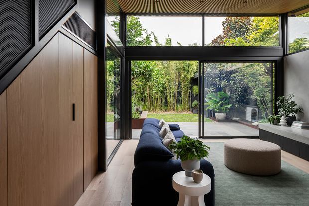 A contemporary living room with floor-to-ceiling black-framed glazing opening onto a lush garden. The room features a dark blue sectional sofa, a round upholstered ottoman, a small white side table, pale timber flooring, dark built-in cabinetry, and a warm slatted timber ceiling. A tall timber door and geometric dark wall detailing are visible along the left side.
