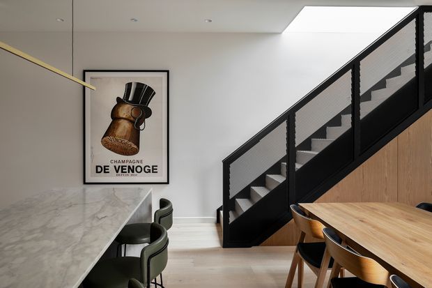 A contemporary open-plan kitchen and dining area beneath a stairway. The space features a large veined stone island with dark green upholstered stools, a timber dining table with matching chairs, pale timber flooring, white walls, and a black-framed stair balustrade with mesh infill. A large skylight brightens the room, while a framed vintage-style champagne poster adds a decorative focal point.