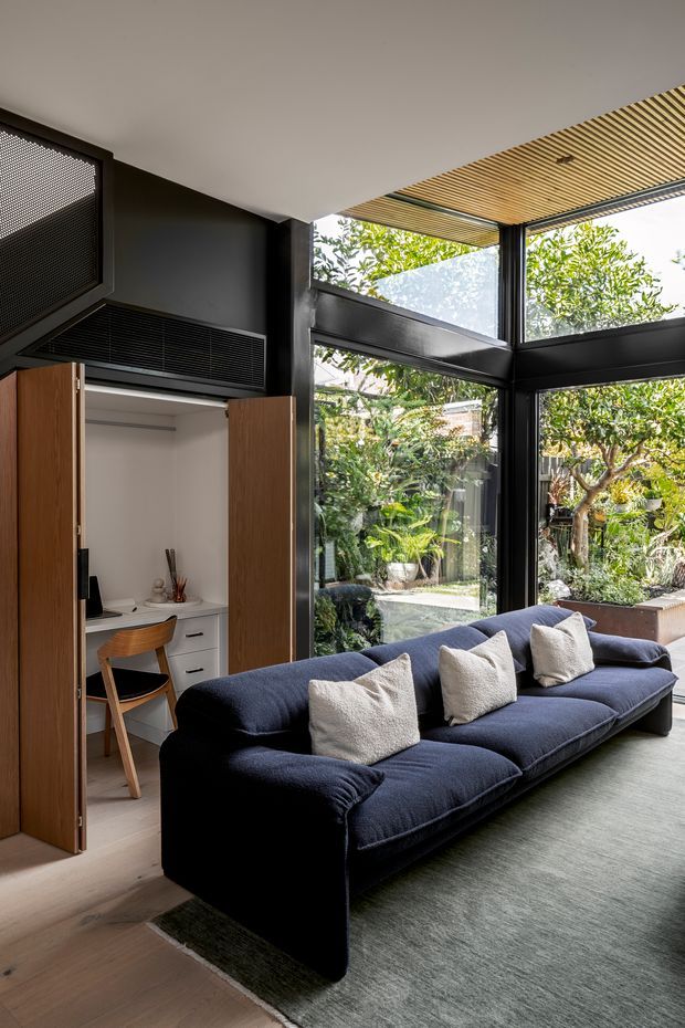 A refined contemporary living room with a large deep navy sectional sofa and neutral cushions, positioned beside full-height black-framed glazing overlooking a leafy courtyard garden. The room includes a compact built-in home office concealed behind timber cabinet doors, dark built-in joinery, pale timber flooring, and a slatted timber ceiling feature extending toward the glazed corner.