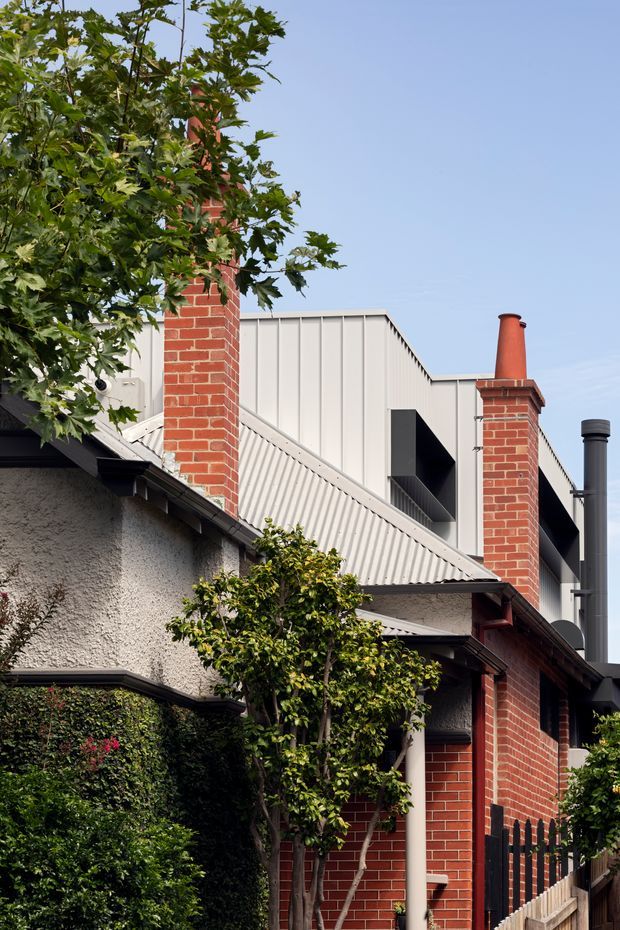 Vertical exterior view of a residential property showing an older red-brick structure with multiple brick chimneys, a textured rendered wall, corrugated metal pitched roofing, and a taller contemporary addition clad in pale vertical metal panels behind it. Mature leafy planting partially screens the building, creating a layered heritage-and-modern streetscape.