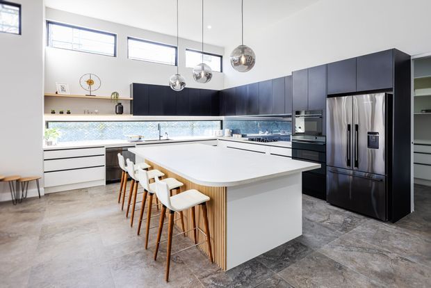 Spacious contemporary kitchen with a large white island, integrated seating, dark charcoal cabinetry, stainless-steel appliances, a patterned tile splashback, and polished spherical pendant lights. High horizontal windows provide abundant natural light, while timber slats add warmth to the island base.