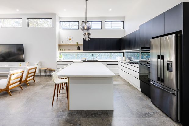 Bright contemporary open-plan kitchen with a large white island, extensive matte black cabinetry, integrated stainless-steel appliances, and a grey stone-look tiled floor. A dining area and living room with television are visible to the left. High horizontal windows and clustered pendant lights add natural light and visual interest.