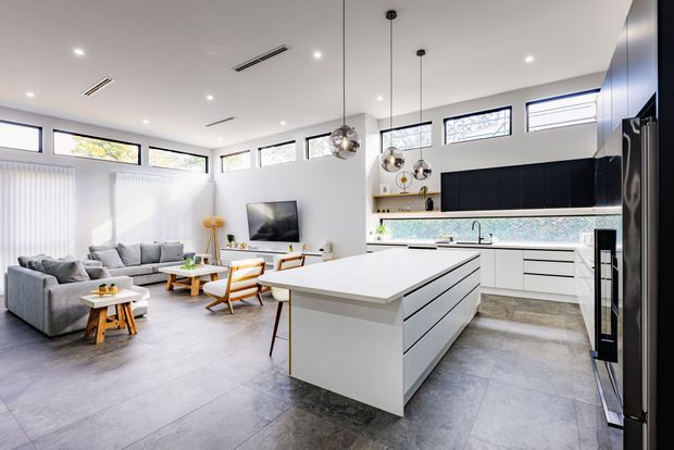 Bright open-plan contemporary kitchen and living area with a large white island, white cabinetry, dark feature joinery, stone-look flooring, pendant lights, high-level clerestory windows, and a grey sectional sofa.