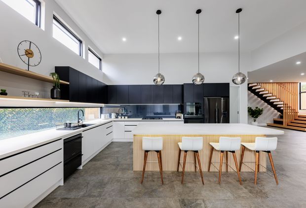 A bright, contemporary open-plan kitchen with white lower cabinetry, dark navy upper cabinets, a long island with integrated seating, and three spherical pendant lights. The kitchen features a textured blue-grey splashback, dark stone-look flooring, stainless-steel appliances, and a timber-slatted island face. A timber staircase and high clerestory windows are visible in the background.