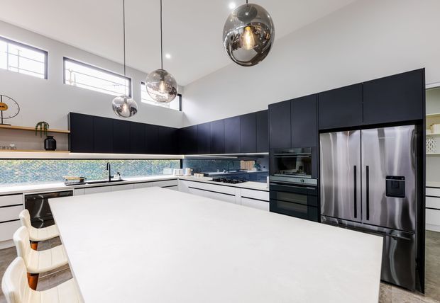 Contemporary open-plan kitchen with a large white island, dark navy-black cabinetry, integrated black appliances, and a stainless-steel French-door refrigerator. A blue mosaic tile splashback runs along the rear benchtop, while polished spherical pendant lights hang beneath a high white ceiling with clerestory windows.