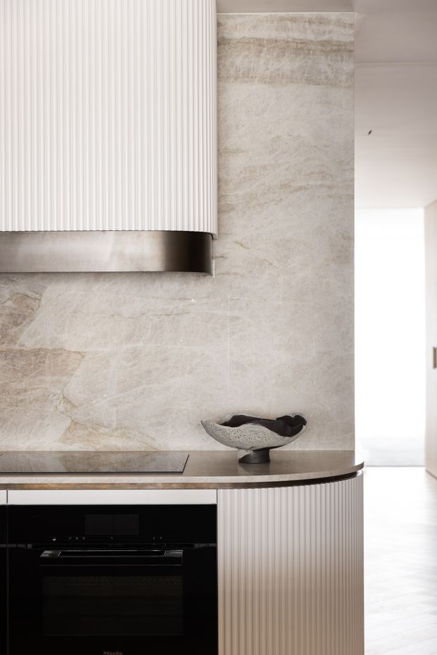 Close-up view of a refined contemporary kitchen featuring a vertically fluted white integrated cabinet and curved peninsula, a dark stone benchtop, a black built-in oven and cooktop, and a large pale veined stone splashback extending up the wall. A sculptural dark bowl sits on the benchtop, with a bright hallway visible beyond.