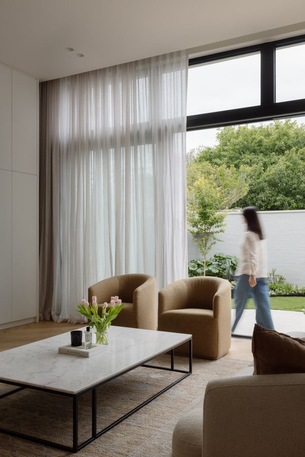 A refined contemporary living room with a neutral palette, featuring two rounded upholstered armchairs and a matching sofa around a rectangular marble-top coffee table with a slim dark metal frame. Full-height sheer curtains frame large black-framed windows and a garden courtyard beyond. Built-in white cabinetry, warm timber flooring, and a textured area rug complete the calm, softly lit interior.