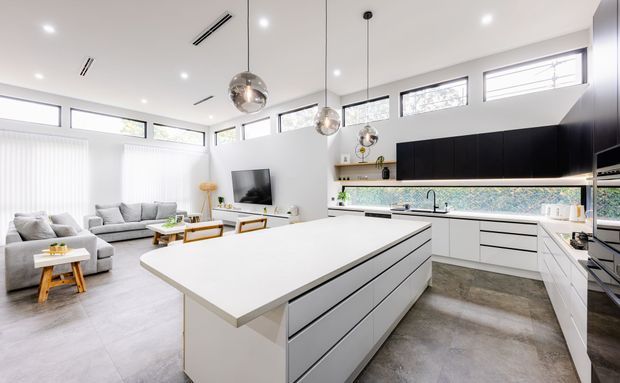 Bright, open-plan contemporary kitchen and living area with a large white island, extensive black-and-white cabinetry, integrated appliances, concrete-look flooring, high clerestory windows, and pendant lighting. A relaxed living room with grey sofas is visible beyond the kitchen.