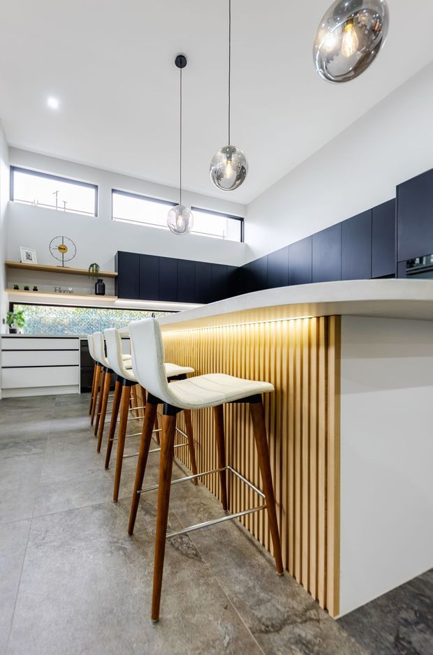 Bright contemporary kitchen with a long island breakfast bar, white upholstered timber stools, dark navy flat-panel cabinetry, and a timber slatted island face with integrated warm lighting. The space has a high ceiling, clerestory windows, polished globe pendant lights, open shelving, and large-format grey stone-look flooring.