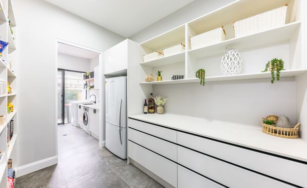 Bright, contemporary laundry and utility room with extensive white built-in cabinetry, open display shelving, a broad white benchtop, integrated refrigerator, and washing machine. The room has light grey tiled flooring, white walls and ceiling, slim black drawer lines, and a connecting doorway to a utility area with a sink and laundry appliances. Minimal decorative accessories add a clean, organised feel.