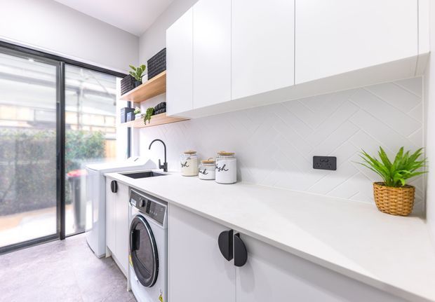 Bright, compact contemporary laundry with white cabinetry, a long pale benchtop, integrated washing machine, sink, black tapware and hardware, and a white geometric tiled splashback. Open timber shelving with small plants and storage jars sits beside full-height glazing and a sliding glass door.