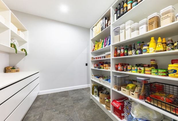 Bright, spacious walk-in pantry with extensive open white shelving, built-in white drawers, organised food containers, bottles, cans and grocery supplies. The room has a white painted finish, recessed ceiling lighting and large-format grey stone-look floor tiles.