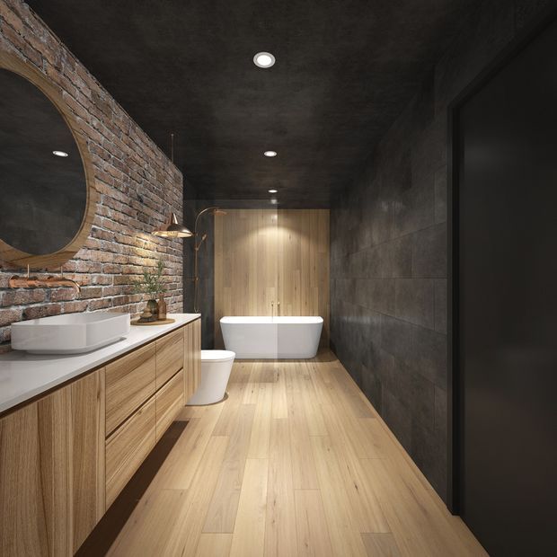 A sophisticated industrial-style bathroom with a freestanding white bathtub, wall-mounted toilet, vessel sink, and timber vanity. The space combines exposed red brick, dark concrete-look wall and ceiling finishes, warm timber flooring and cabinetry, black-framed glazing or door elements, and warm recessed and pendant lighting.