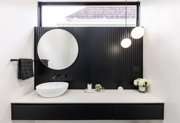 Contemporary bathroom vanity with a long white benchtop, matte black floating cabinetry, black vertical fluted feature wall, round mirror, vessel basin, wall-mounted tapware, and spherical pendant lights. A high horizontal window brings in daylight, with minimal monochrome styling and small plants.