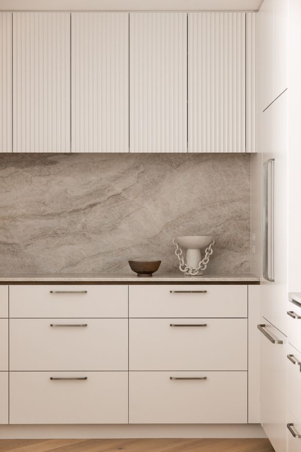 A close-up view of a refined contemporary kitchen, featuring full-height pale ribbed cabinetry, smooth light cabinetry below, long slim metal handles, a large taupe-grey stone backsplash and benchtop, and warm timber flooring. Decorative ceramic vessels sit on the benchtop, while a tall integrated appliance cabinet is visible at right.
