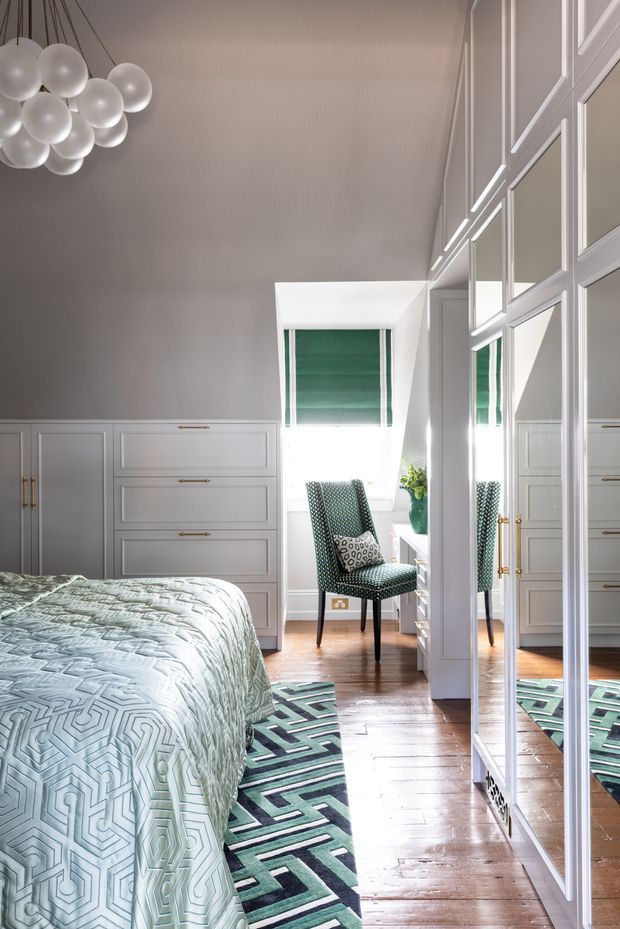A bright, refined bedroom with pale grey built-in cabinetry, mirrored wardrobe doors, and timber floorboards. A patterned green-and-white bedspread and geometric rug add colour, while a dark patterned occasional chair sits by the window with a green roller blind. White globe pendant lighting and decorative wall moulding contribute to the polished, classic interior.