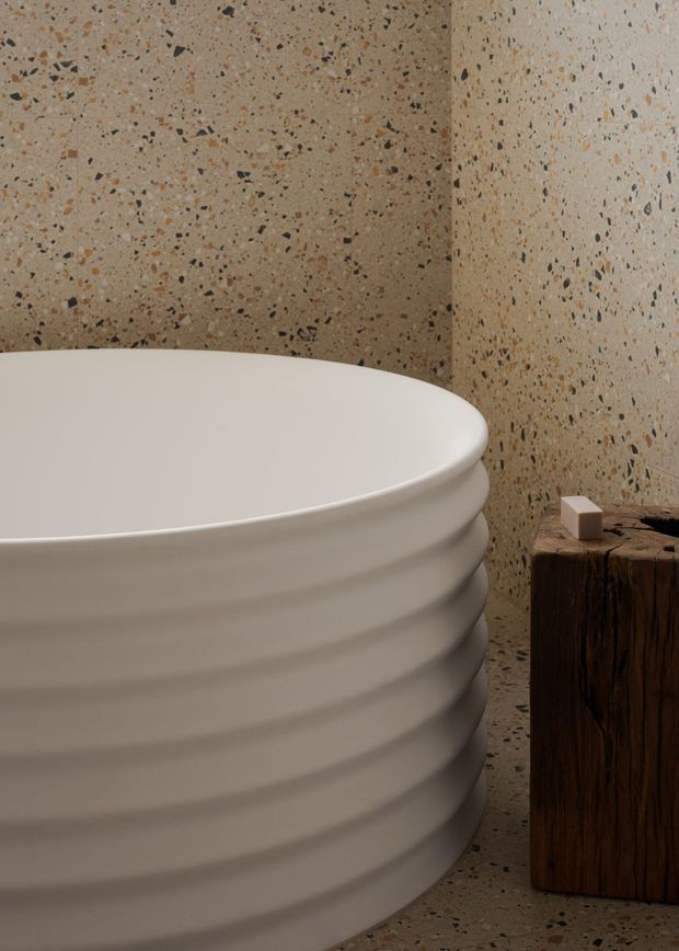 Close-up of a sculptural freestanding white bathtub with horizontal ribbed detailing, set against warm speckled terrazzo walls and flooring. A dark natural timber pedestal or side table sits beside the tub, creating a restrained, tactile bathroom composition.