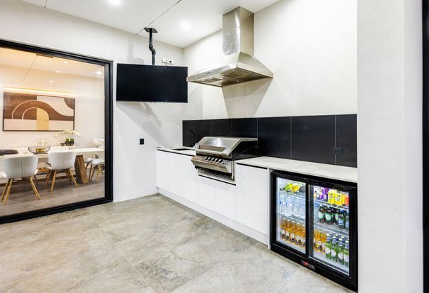 A compact contemporary commercial-style kitchen with white lower cabinetry, a black tiled splashback, stainless-steel rangehood and cooking equipment, and a large glass-front drinks refrigerator. The space has a polished concrete-look floor and opens visually to a dining area with a table, chairs, and framed artwork.