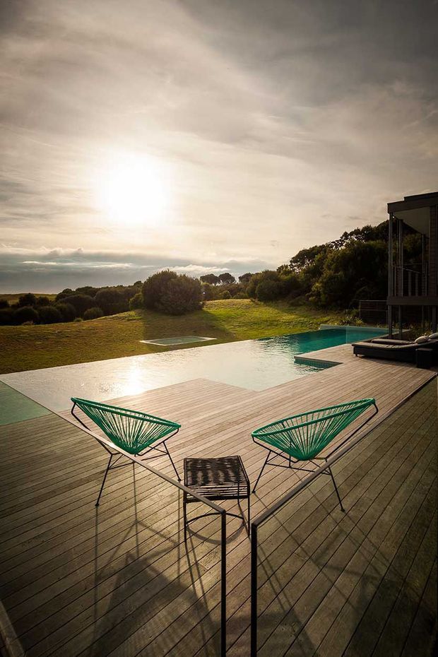 A wide timber deck overlooks an infinity pool and landscaped grassy terrain at sunset. Two green wire-frame lounge chairs and a small matching table occupy the foreground, while a modern building with dark vertical elements is partially visible at right. The scene has a warm, tranquil outdoor resort-like atmosphere.