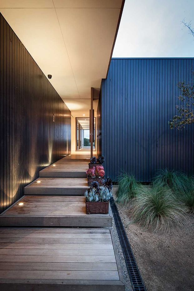 A narrow, covered exterior walkway leading toward a modern home entrance, with broad timber decking steps, dark vertical cladding walls, a blue-grey vertically ribbed facade, recessed floor lighting, and a linear drainage channel beside landscaped ornamental grasses and potted plants. The sheltered corridor creates a strong framed perspective toward glazed doors at the far end.
