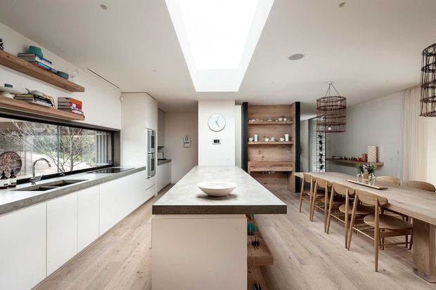 A bright, open-plan contemporary kitchen and dining area with white flat-panel cabinetry, a long stone island, pale timber flooring, timber open shelving, and a large dining table. A wide window runs along the kitchen bench, while a large linear skylight brings abundant natural light into the space. Warm timber accents and pendant-style wire lights create a refined, inviting atmosphere.