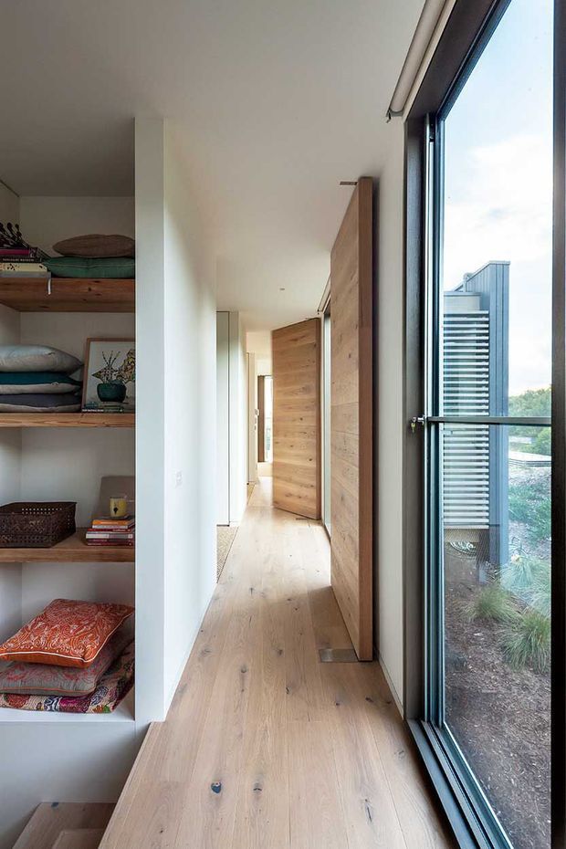 A bright, narrow residential hallway with pale timber flooring and matching timber sliding or pivot doors along one side. Built-in open shelving displays folded linens and small decorative items, while full-height dark-framed glazing provides views to an adjacent exterior landscape and neighbouring building. The interior is simple, warm, and contemporary with clean white walls and minimal detailing.