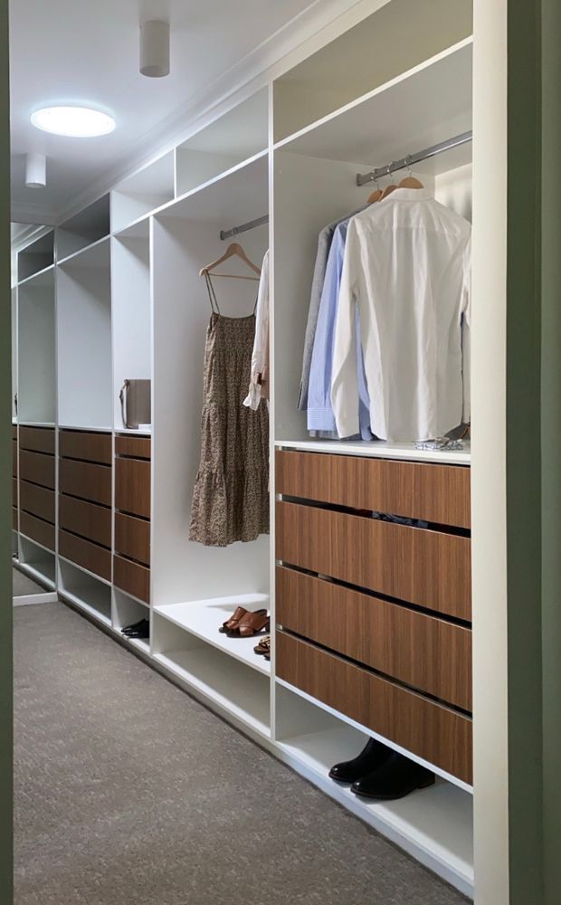 A well-organised walk-in wardrobe with full-height white open shelving, hanging rails, and integrated dark timber drawer cabinetry. Clothes and shoes are neatly arranged throughout, with mirrored panels along the left side and recessed ceiling lighting. The room has a neutral, refined finish and a carpeted floor.