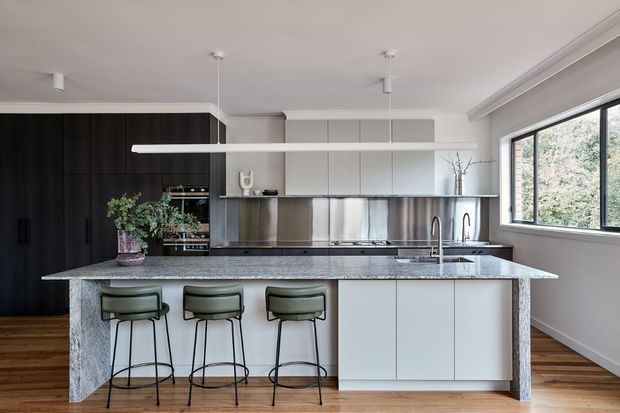 A bright contemporary kitchen with a large stone island and three bar stools, dark full-height cabinetry on the left, white handleless cabinetry at the rear, stainless-steel appliances, timber flooring, and large windows overlooking greenery. A slim suspended white shelf runs above the island.