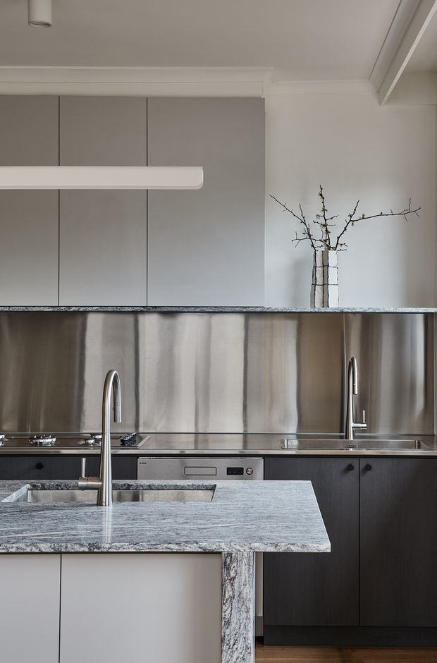 A refined contemporary kitchen viewed across a stone island. The space features dark timber cabinetry, a dramatic stainless-steel splashback, pale grey upper cabinets, integrated appliances, twin undermount sinks with tall gooseneck taps, and a heavily veined grey stone island benchtop. A simple branch arrangement and linear pendant light add restrained decorative detail.