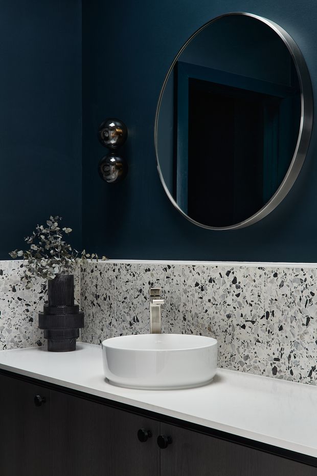 A moody, refined bathroom vanity vignette with a white vessel basin on a white countertop, dark timber cabinetry, a terrazzo-look splashback, deep navy walls, a chrome tap, and a large oval mirror. Decorative black wall lights and a dark ribbed vase add contrast.