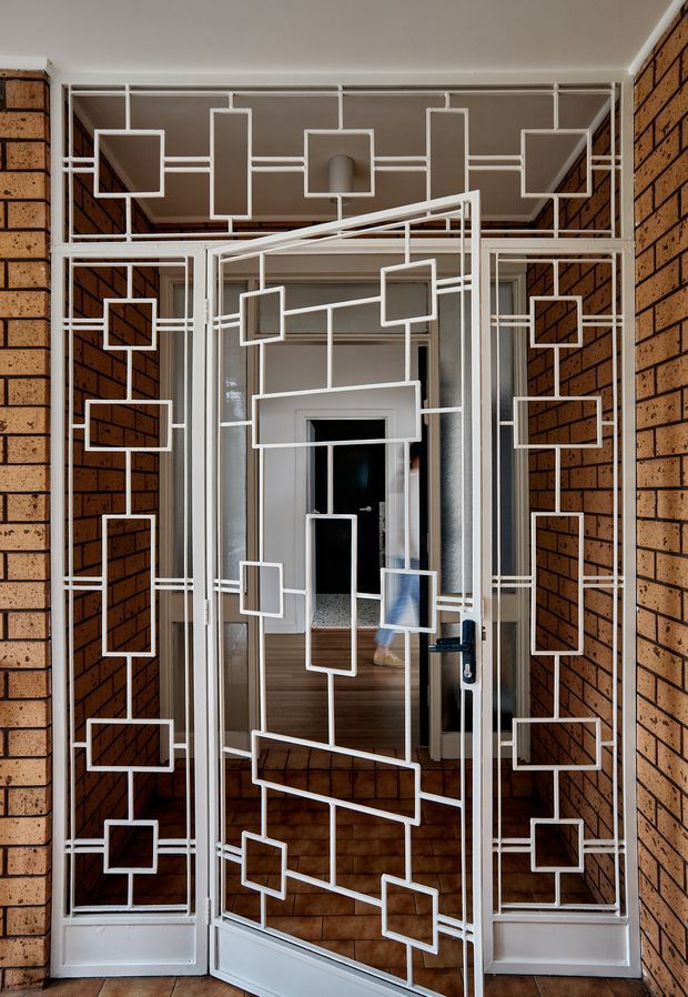 Interior view of a residential entryway featuring a distinctive white-painted metal security screen door and matching sidelights decorated with an abstract geometric grid of squares and rectangles. Exposed warm-toned brick walls frame the entrance, while a glazed inner door and timber flooring are visible beyond.