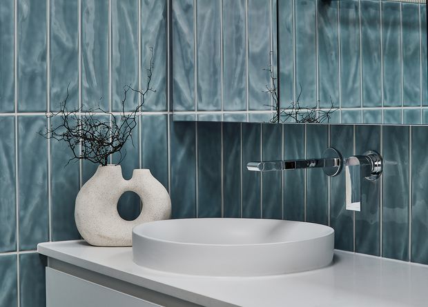 Close-up of a contemporary bathroom vanity with a round white countertop basin on a pale benchtop, wall-mounted chrome tapware, and vertically arranged glossy blue-grey tiles. A sculptural neutral vase with bare branches provides subtle decor.