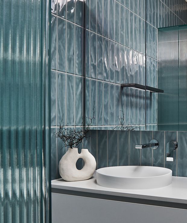 Close-up of a refined contemporary bathroom vanity. A white vessel basin sits on a pale stone or solid-surface benchtop above grey cabinetry, with wall-mounted chrome tapware and a dark-framed glass shelf. The walls combine glossy blue-grey rectangular tiles with vertically ribbed translucent glass or tile panels, creating a cool, polished and textural finish. Minimal decorative styling includes a sculptural white vase with bare branches.