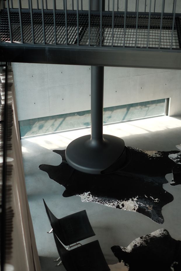 A dramatic double-height interior with a dark cylindrical structural column supporting a mezzanine or upper-level walkway. The space features smooth concrete walls and flooring, extensive metal balustrading and grating, strong sunlight, and black-and-white cowhide rugs beneath dark modern seating. The composition highlights industrial materials, vertical volume, and graphic contrast.
