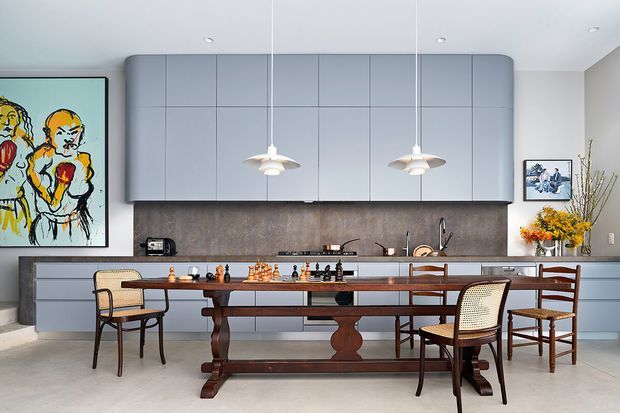 A refined open-plan kitchen and dining area with cool grey-blue cabinetry, a dark textured splashback, pale stone flooring, and a long dark timber dining table surrounded by mixed classic and woven chairs. Two pendant lights hang above the table, while artwork and floral arrangements add colourful accents.