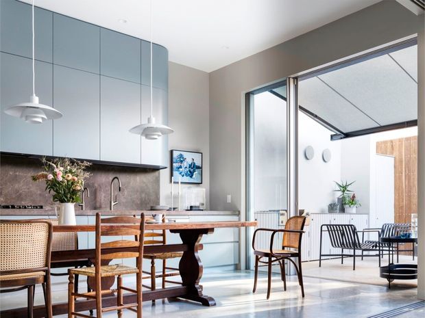Bright, refined open-plan kitchen and dining area with pale blue-grey curved cabinetry, a dark stone backsplash, timber dining table and vintage-style timber and cane chairs. Large sliding glass doors open to a covered outdoor courtyard or terrace with additional seating, creating a strong indoor-outdoor connection. Pendant lights, framed artwork and indoor plants add warmth to the contemporary space.